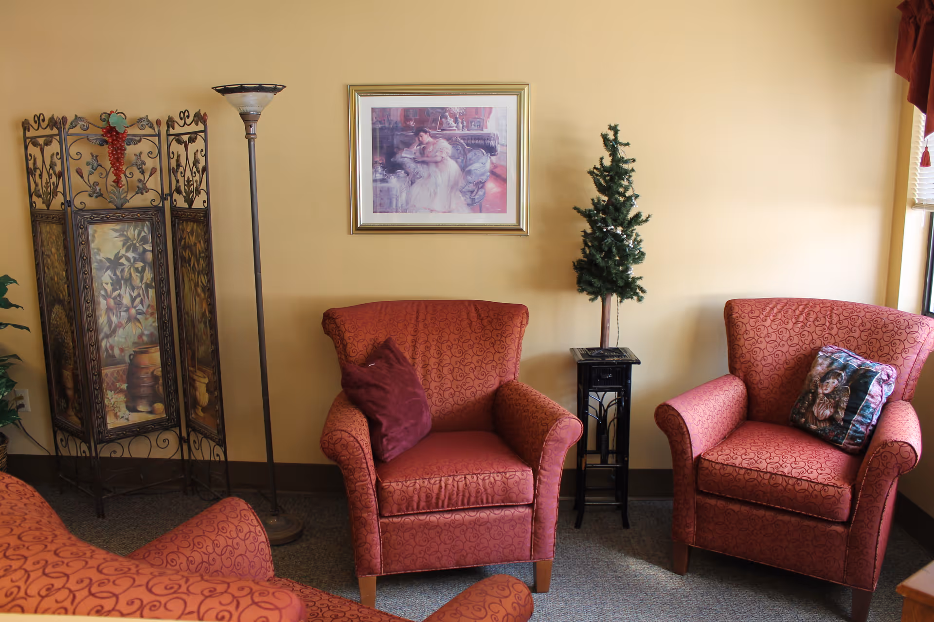 A cozy sitting area with three red patterned armchairs, each with a decorative pillow. There is a small artificial Christmas tree on a black stand between two of the chairs. A tall floor lamp and a decorative folding screen with a grapevine design are against a beige wall. A framed painting hangs above the center chair.