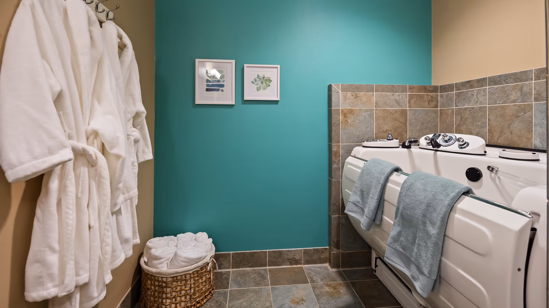 A bathroom with a walk-in bathtub featuring control knobs and two light blue towels hanging on the side. On the left wall, there are three white bathrobes hanging on hooks. Below the robes, a wicker basket holds several rolled white towels. The walls are painted teal and beige, with tiled flooring and partial tiled walls around the bathtub. Two framed botanical prints hang on the teal wall.