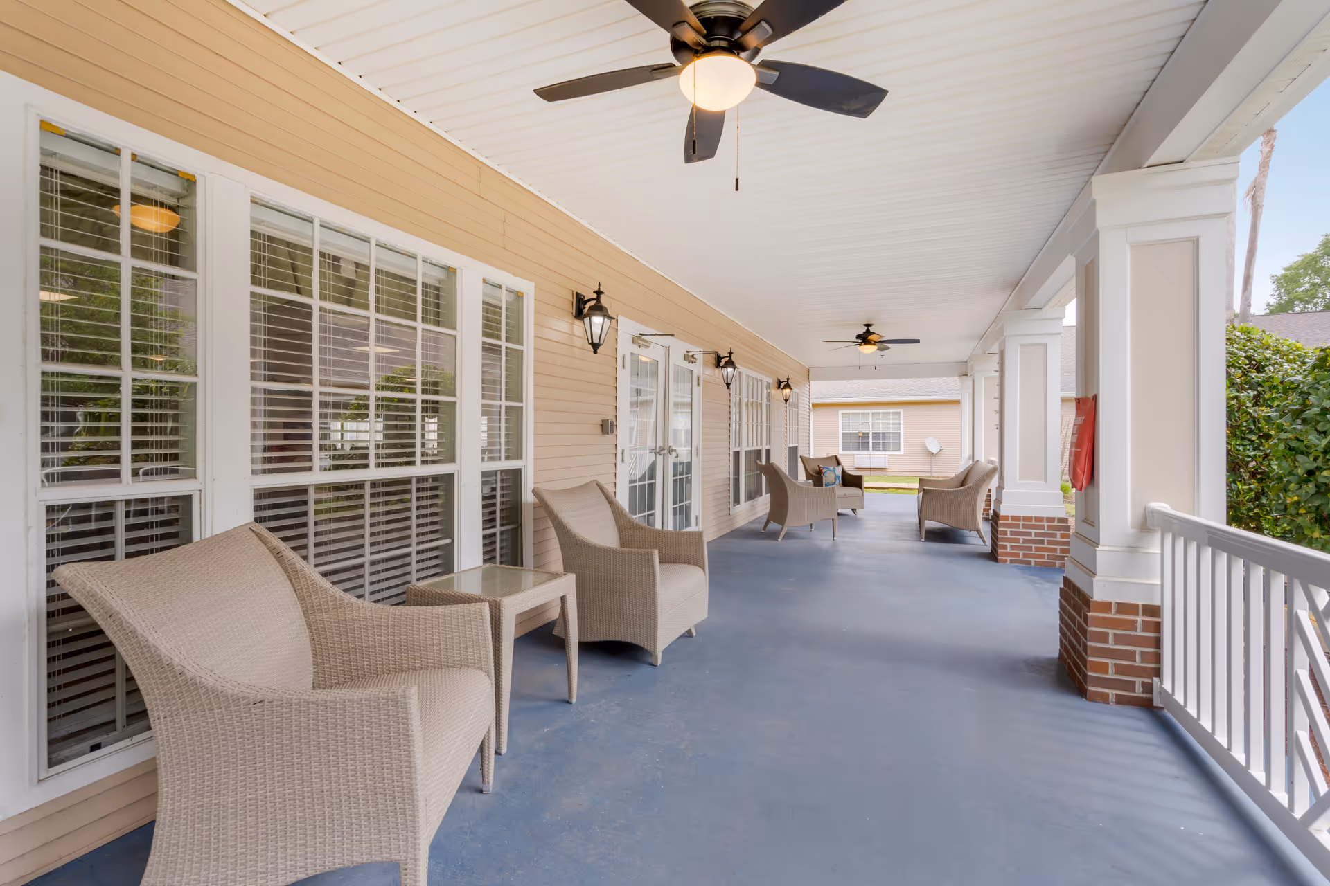 Covered porch with wicker seating, ceiling fans, and windows along a building exterior.