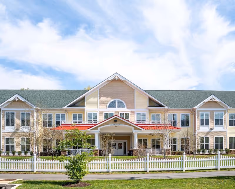 Front exterior of a two-story yellow senior living building with a central covered entrance, white picket fence, and green roof under a blue sky.