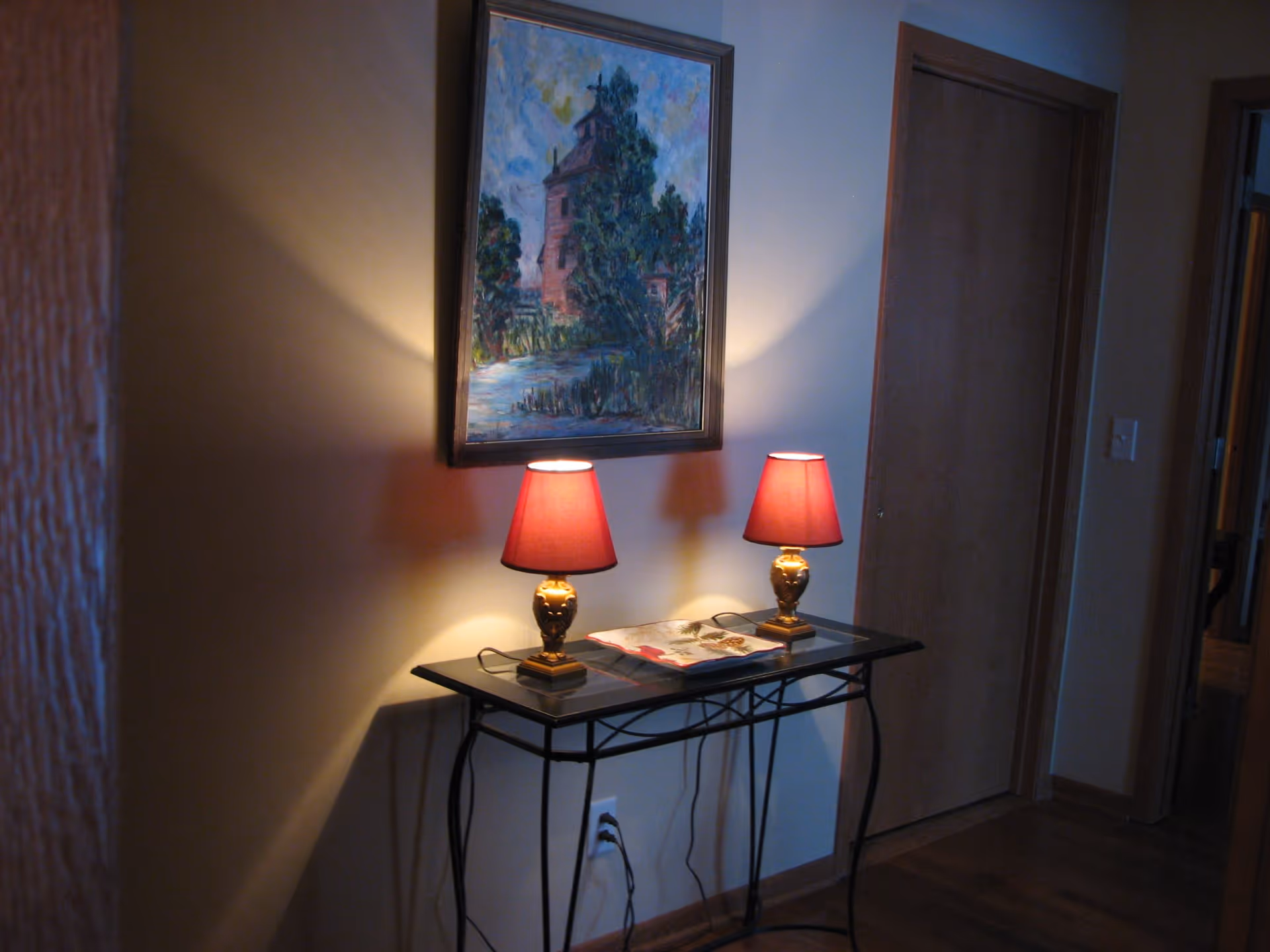 A hallway corner with a narrow black metal table holding two small lamps with red shades and a decorative plate. Above the table hangs a framed painting of a building surrounded by trees. The walls are light-colored, and there is a wooden door to the right.