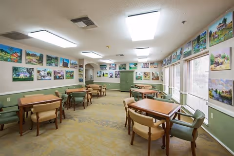 A well-lit dining room with multiple wooden tables and cushioned chairs arranged neatly. The walls are decorated with numerous colorful paintings depicting various outdoor scenes. Large windows on one side allow natural light to enter the room.