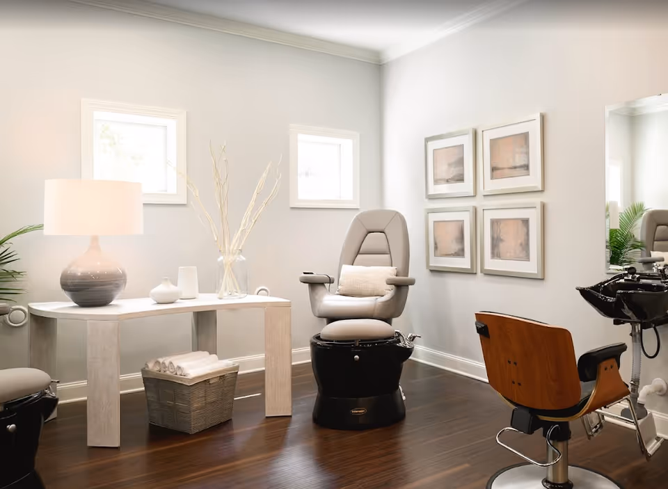 A bright modern interior salon room with reclining shampoo chairs, a wooden styling chair, a console table with a lamp and decor, and framed artwork on the wall.
