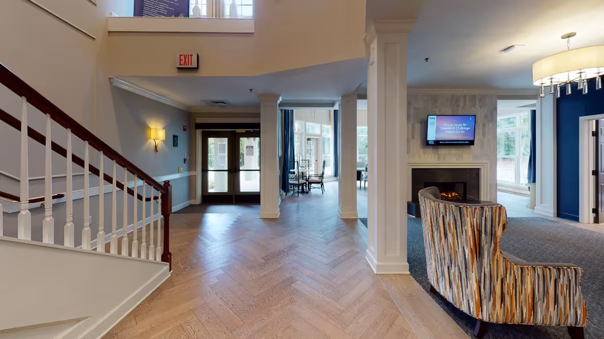 Interior view of a senior living facility lobby area with a staircase on the left, double glass doors straight ahead, and a seating area with a fireplace and a wall-mounted TV on the right. The floor is light wood with a herringbone pattern, and there are large windows letting in natural light.