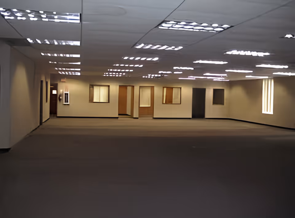 A large empty interior room with beige walls, multiple doorways and windows, and a ceiling with many fluorescent lights.