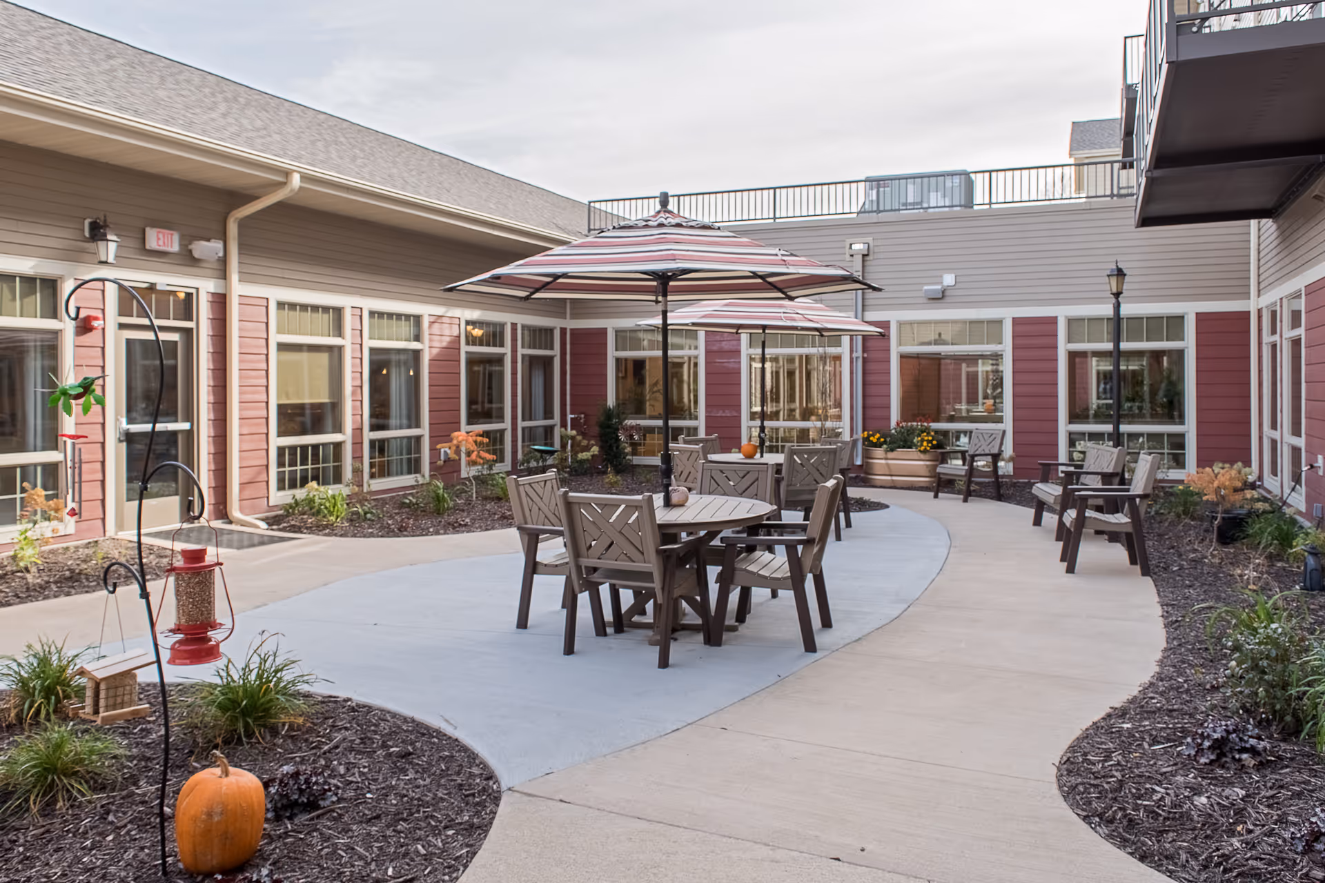 Outdoor courtyard area at a senior living facility with round tables and chairs under striped umbrellas, surrounded by a walkway and garden beds with plants and pumpkins. The building has large windows and beige and red siding.