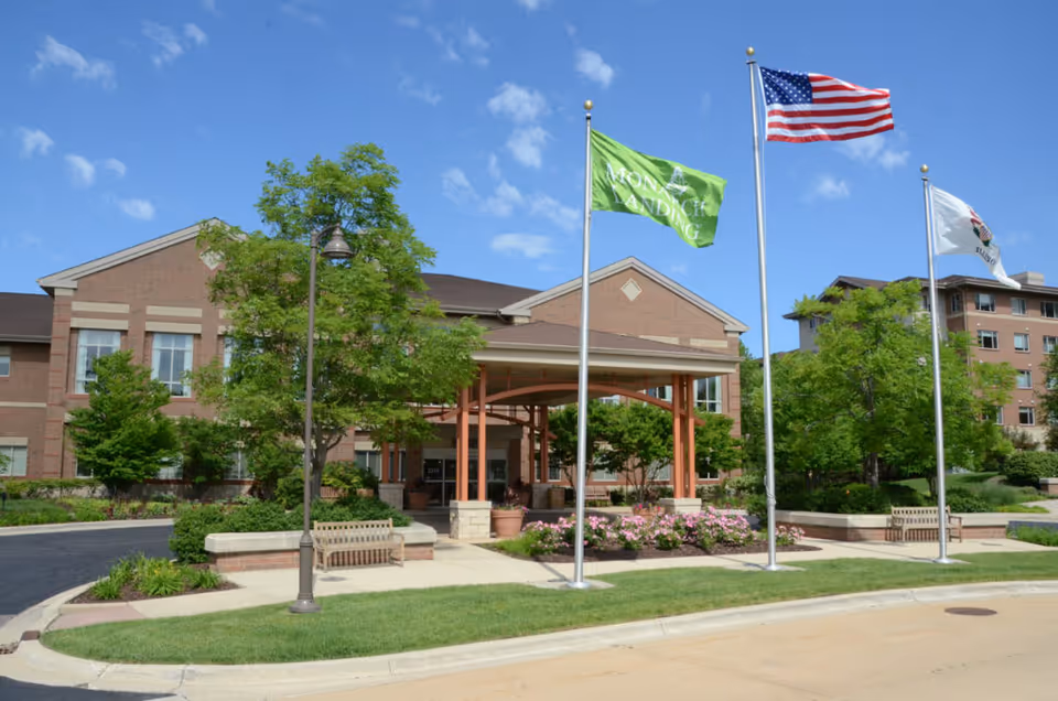 Exterior view of Monarch Landing senior living facility with a brick building, three flagpoles displaying the Monarch Landing flag, the American flag, and another flag, surrounded by green trees, benches, and a clear blue sky.