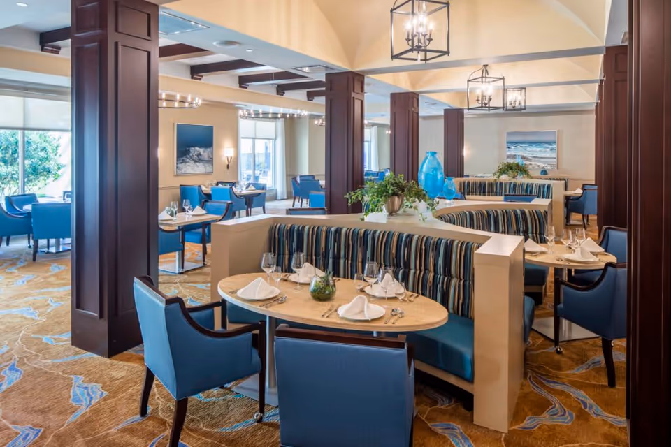 A spacious dining room with multiple tables set for meals, featuring blue upholstered chairs and striped booth seating. The room has large windows allowing natural light, decorative plants on tables, and framed ocean-themed artwork on the walls. The ceiling has exposed beams and hanging lantern-style light fixtures.