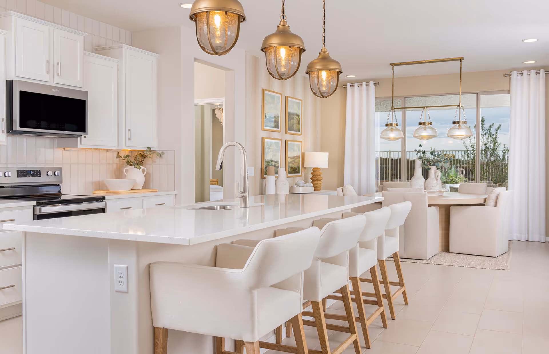 Bright open-concept kitchen with a long white island, upholstered bar stools, brass pendant lights, and a dining area by a large window.