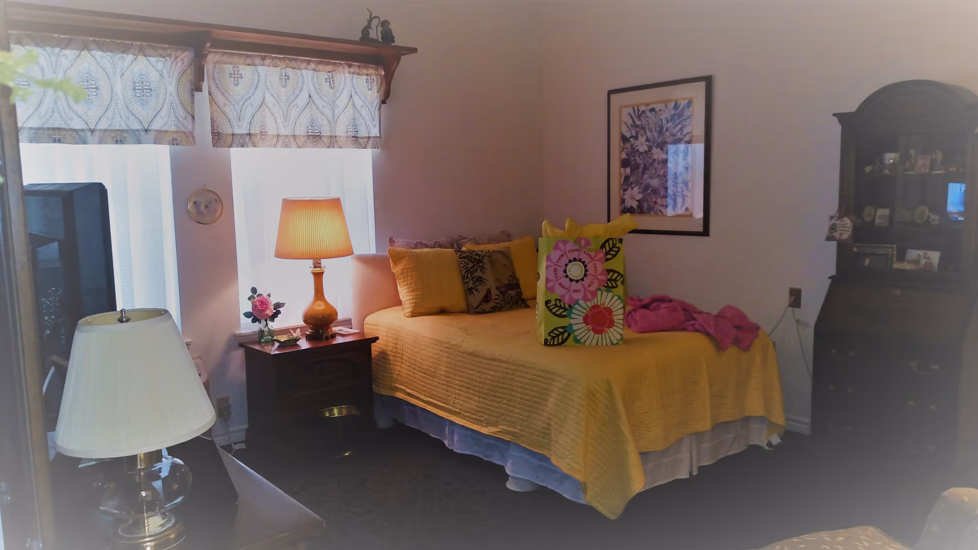 A cozy bedroom with a single bed covered in a yellow quilt and several pillows. A colorful gift bag and a pink blanket are on the bed. There is a wooden nightstand with a lamp and a small vase with a pink flower next to the bed. Two windows with patterned valances let in natural light. A framed floral picture hangs on the wall above the bed. A dark wooden cabinet with glass doors displaying various items is on the right side of the room. Another lamp with a white shade is visible in the foreground.