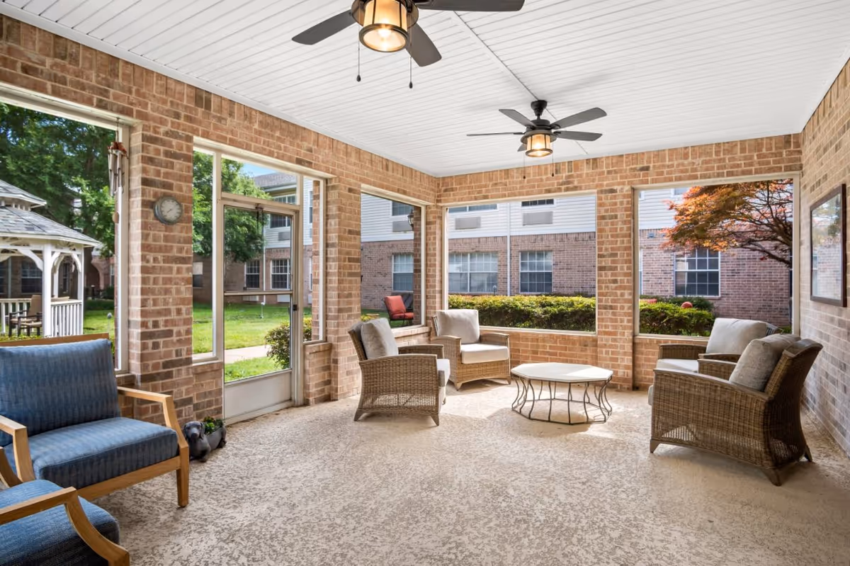 Bright enclosed brick sunroom with wicker chairs, ceiling fans, and windows overlooking a courtyard.