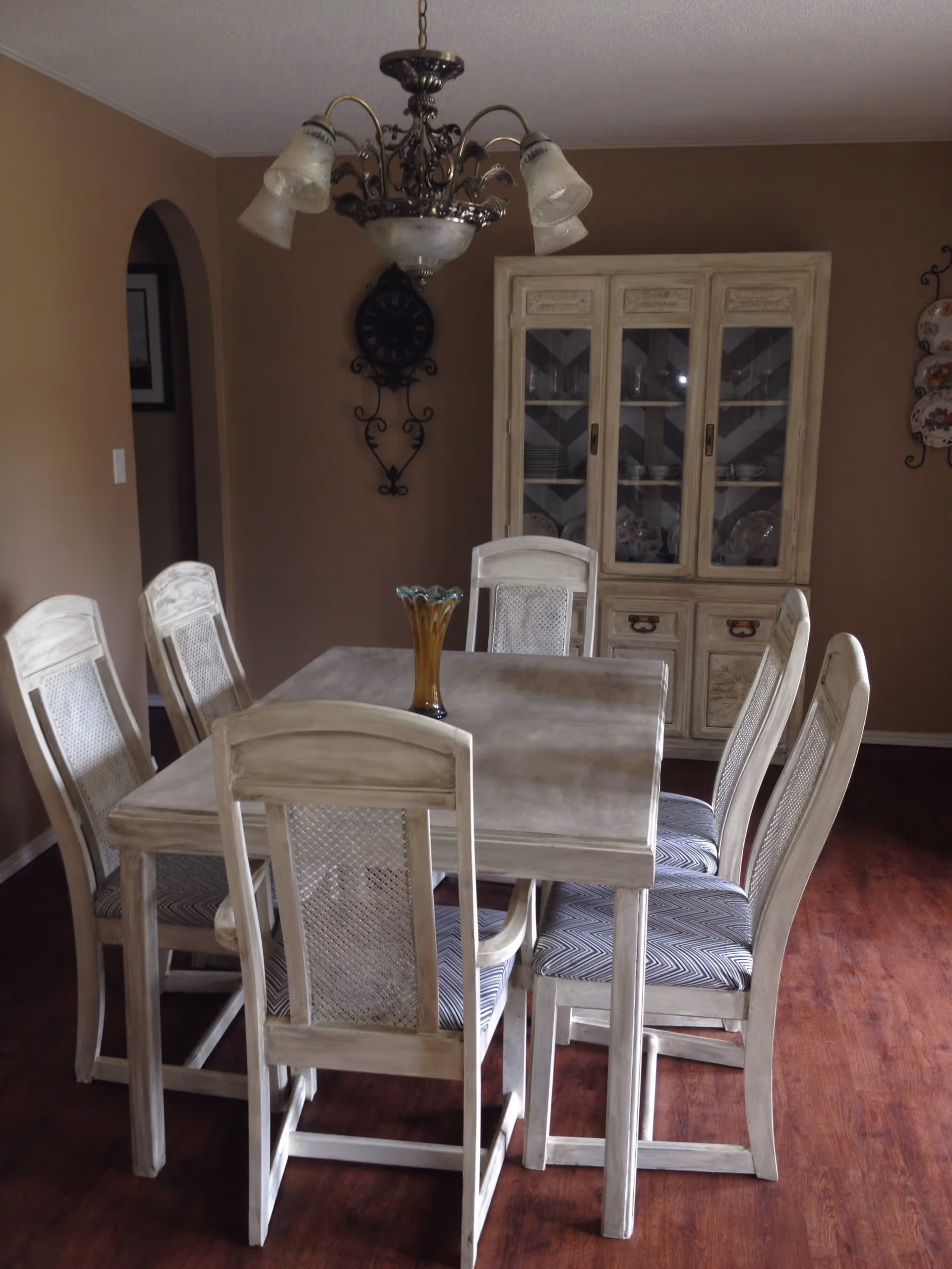 A dining room with a rectangular wooden table surrounded by six matching chairs with patterned cushions. A decorative glass vase is placed in the center of the table. Behind the table is a wooden cabinet with glass doors displaying dishes and glassware. A wall clock with ornate metalwork hangs on the wall, and a chandelier with multiple light fixtures hangs above the table. The room has brown walls and wooden flooring.