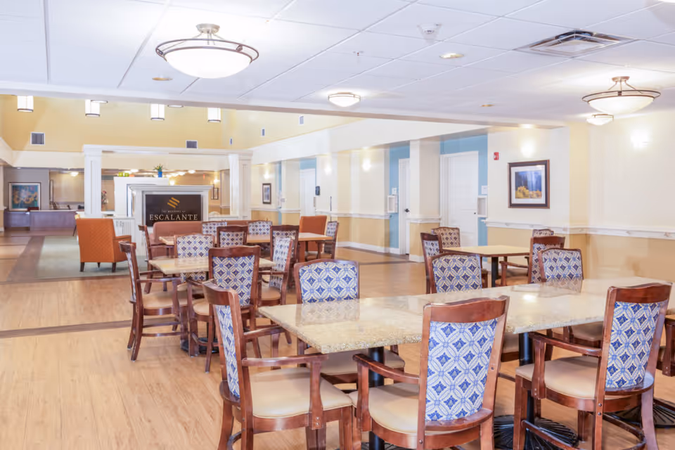 A spacious dining area in a senior living facility with multiple tables and chairs. The chairs have wooden frames with patterned blue and white upholstery. The room has light-colored walls, wood flooring, and ceiling lights. In the background, there is a sign that reads 'The Meadows at Escalante' and some seating areas with orange armchairs.