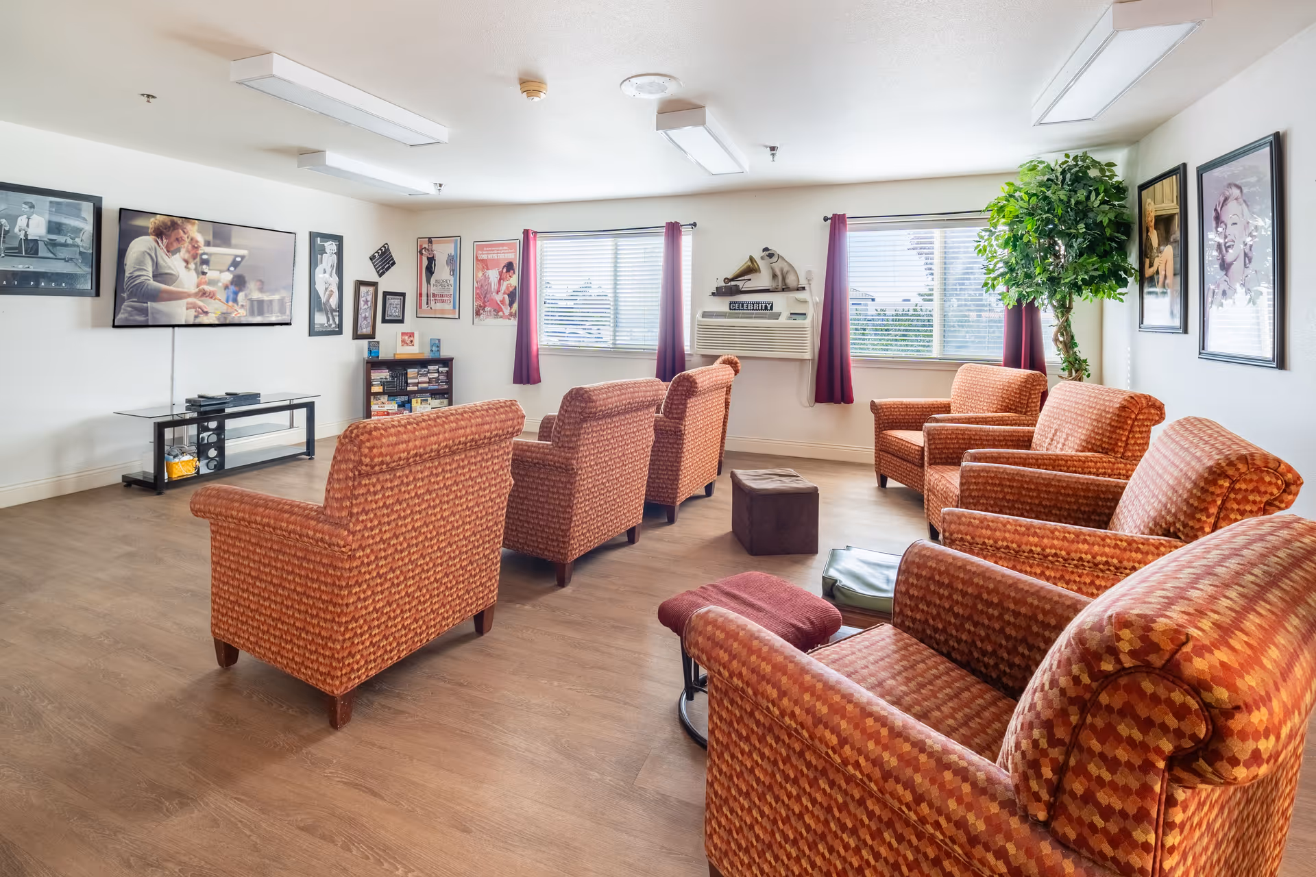 A bright common room with patterned armchairs arranged facing a wall-mounted TV, framed posters, and windows with curtains.