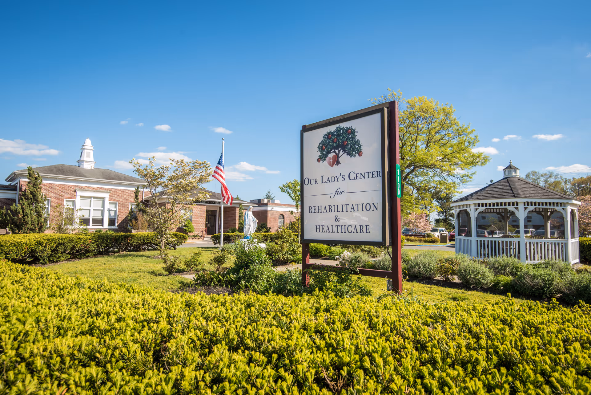 Exterior view of Our Lady's Center for Rehabilitation & Healthcare on a sunny day, showing a large sign with the facility's name, a brick building, an American flag, a statue, and a white gazebo surrounded by green bushes and trees.