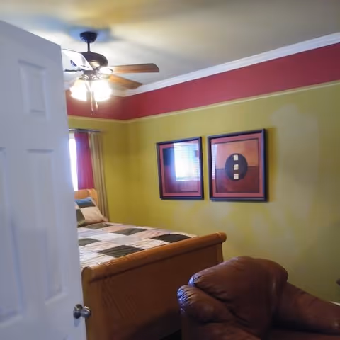 View into a bedroom with a partially open white door. Inside, there is a bed with a multicolored patchwork quilt, a brown leather armchair, two framed abstract artworks on a green wall with a red border near the ceiling, and a ceiling fan with lights.