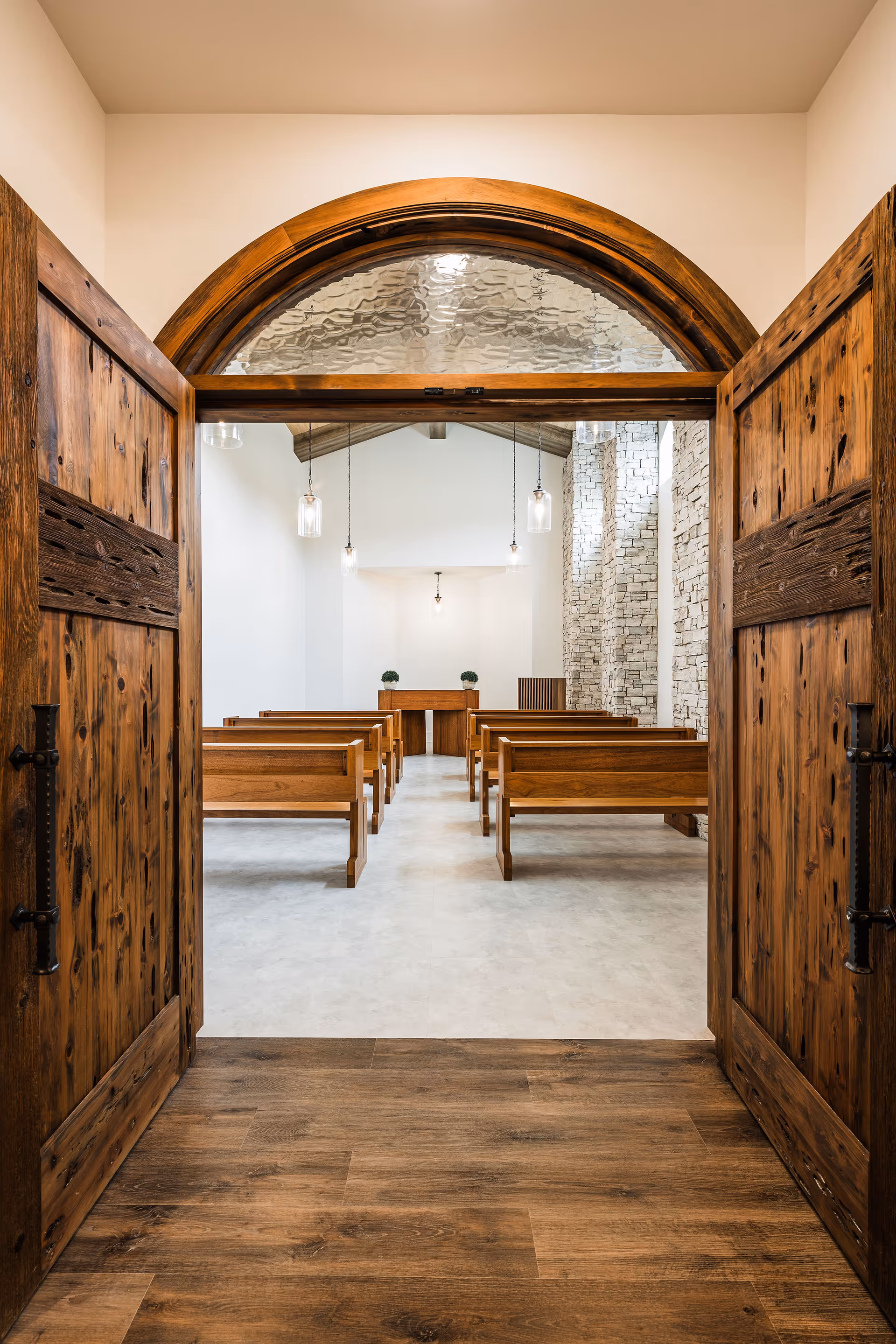 Open rustic wooden double doors leading into a small chapel-like interior with wooden pews, a raised altar, pendant lights, and a stone accent wall.