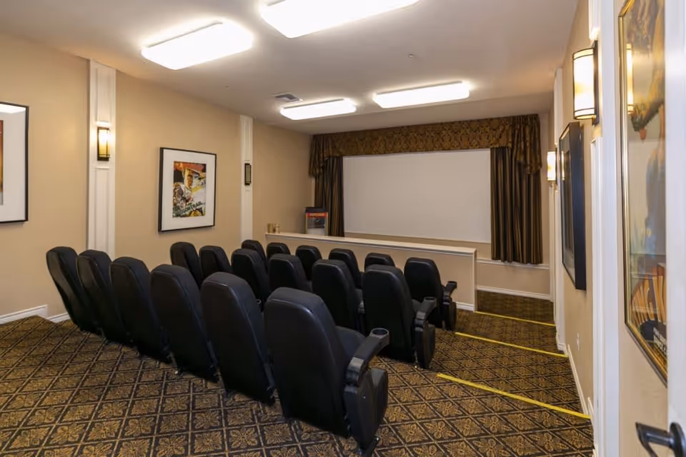 Small theater room with two rows of black cushioned seats facing a large white projection screen. The room has patterned carpet, beige walls with framed movie posters, wall sconces for lighting, and a popcorn machine near the screen.