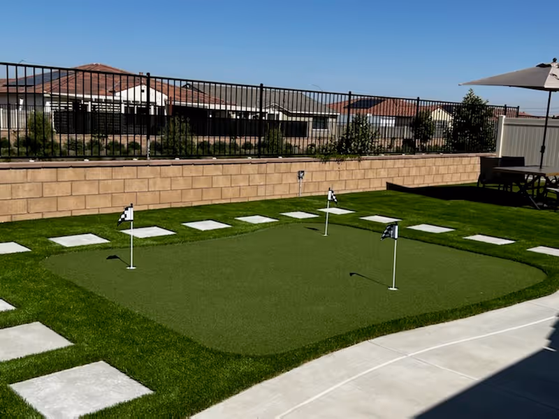 Backyard putting green with three flags, surrounding stepping stones, patio furniture, and a low block wall and fence with neighboring houses beyond.