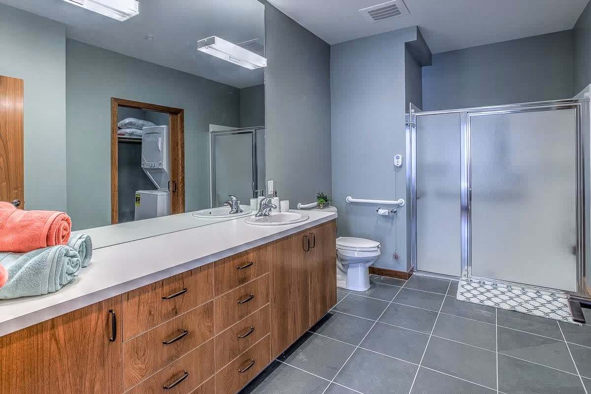 A modern bathroom with gray tiled floor and walls painted in a muted gray tone. There is a long wooden vanity with a white countertop featuring two sinks and chrome faucets. On the countertop, there are neatly folded towels in coral and light blue colors. A large mirror spans the length of the vanity, reflecting a stacked washer and dryer in an adjacent room. To the right, there is a white toilet with a grab bar beside it and a glass-enclosed shower with frosted doors. A small plant and soap dispensers are placed on the countertop near the toilet.