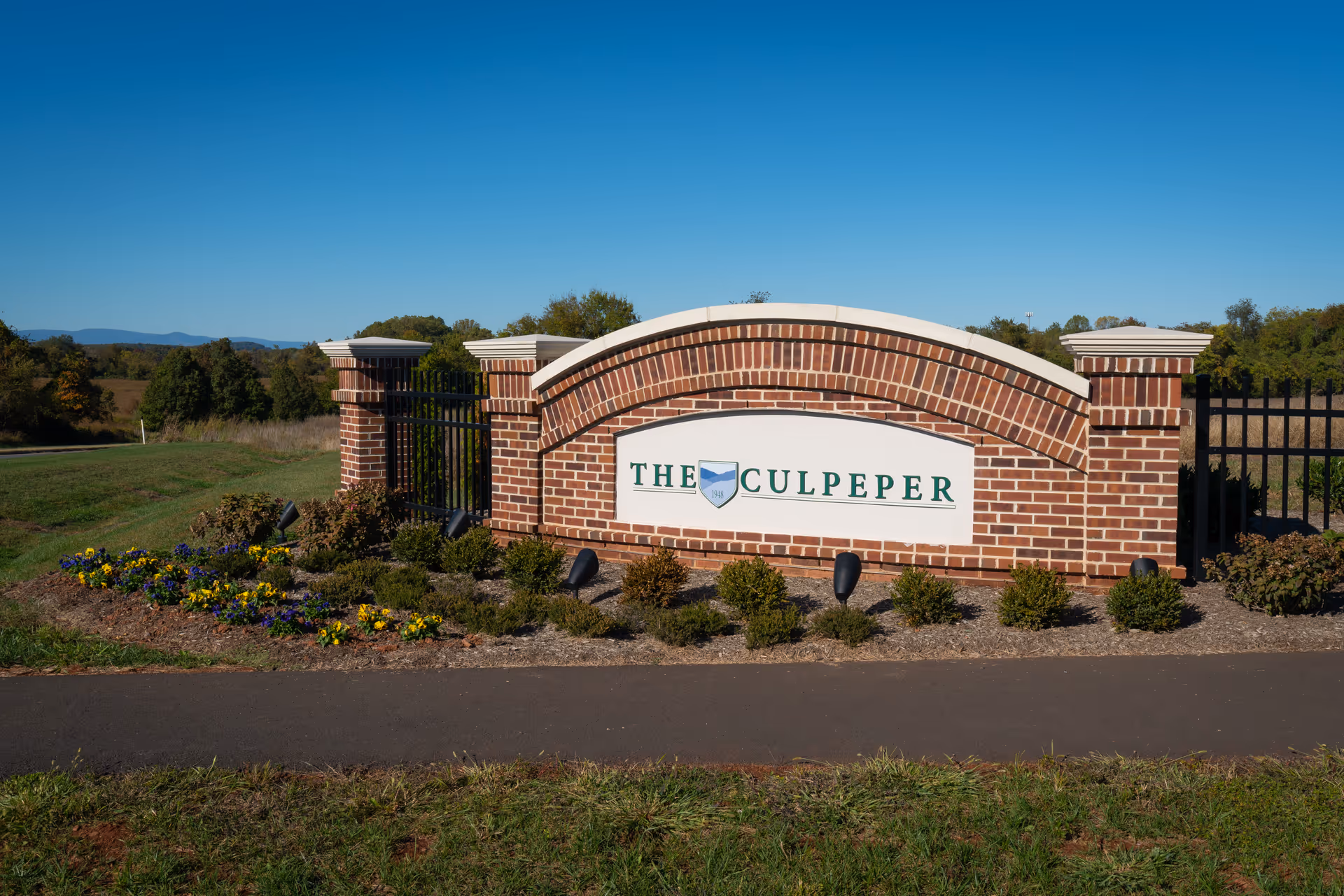 A brick entrance sign for The Culpeper facility, surrounded by a landscaped area with small bushes and colorful flowers, set against a clear blue sky and a natural green landscape in the background.
