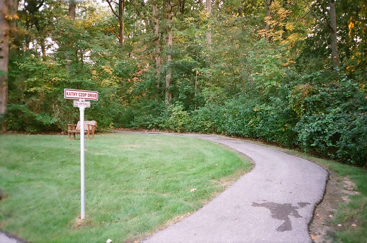 A curved paved pathway surrounded by green grass and dense trees with some autumn foliage. A street sign reads 'Kathy Czop Drive' with a smaller sign below stating 'The Czop Family'. Two wooden benches are placed on the grass near the sign.