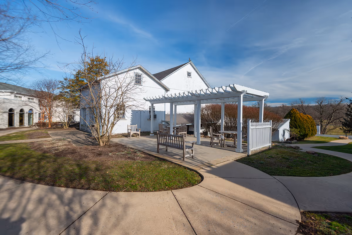 A white pergola and benches on a paved patio beside a white building with walkways and grassy landscaping under a blue sky.