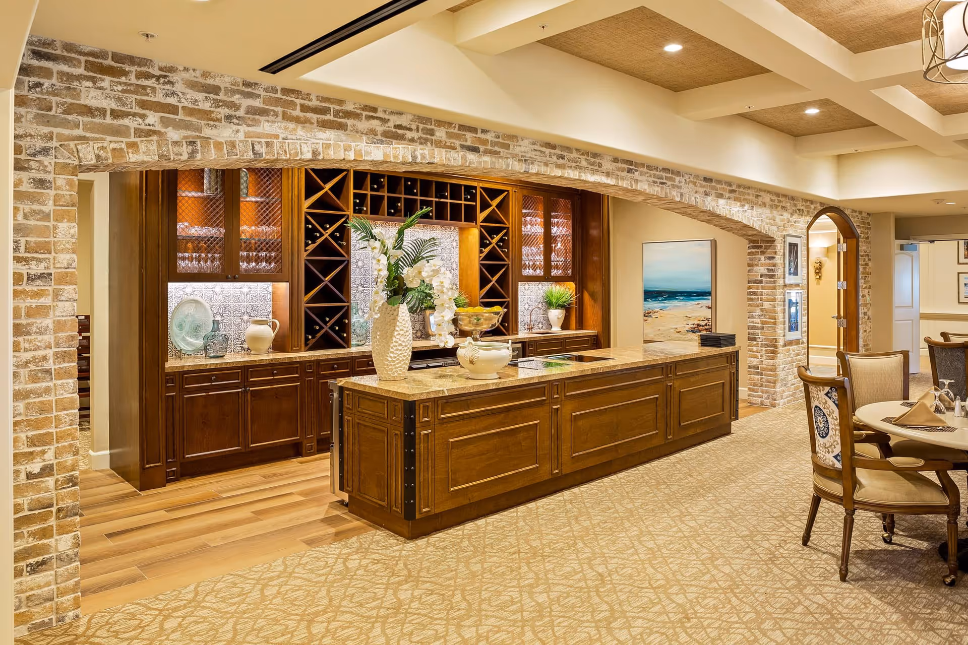 Interior view of a senior living facility dining area featuring a large wooden island with a marble countertop, decorated with vases and plants. Behind the island is a built-in wooden cabinet with wine racks, glassware, and decorative plates, framed by a brick archway. To the right, there is a dining table with upholstered chairs and a painting of a beach scene on the wall.