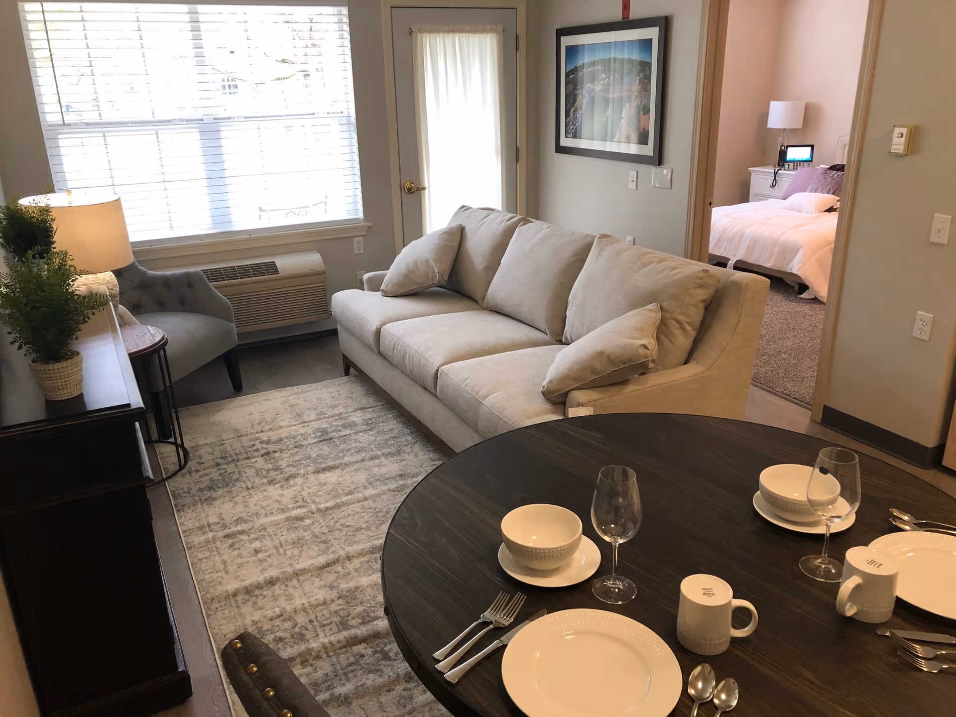 A cozy living room area with a beige sofa and a gray armchair next to a window with blinds. A dark wooden dining table is set with white plates, bowls, mugs, silverware, and wine glasses. A framed picture hangs on the wall, and a bedroom with a white bedspread is visible through an open doorway.