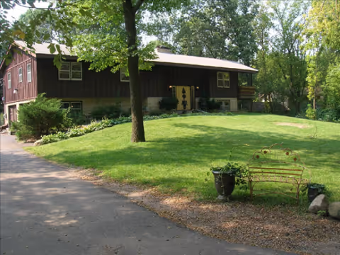 Exterior view of a brown building with a green lawn, bench, and surrounding trees.