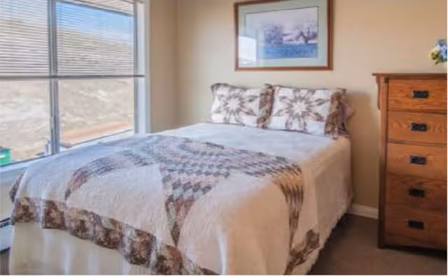 A bedroom with a bed covered in a patterned quilt and matching pillows. There is a wooden dresser with drawers to the right and a framed picture hanging on the wall above the bed. A large window with blinds is on the left side, letting in natural light.