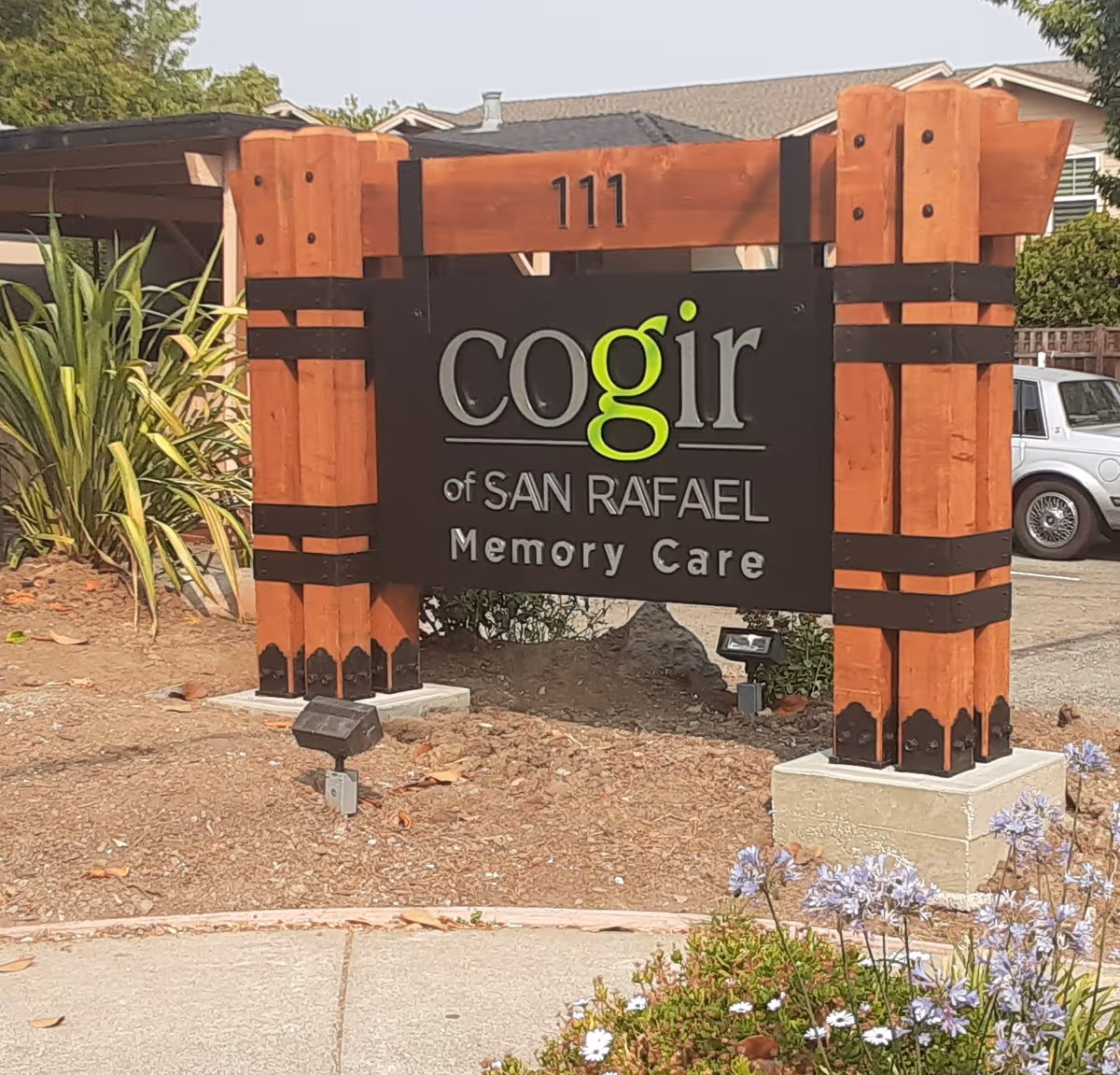 A wooden sign with black and green lettering that reads 'cogir of SAN RAFAEL Memory Care' mounted on a landscaped area with plants and flowers, with a building and a parked car in the background.