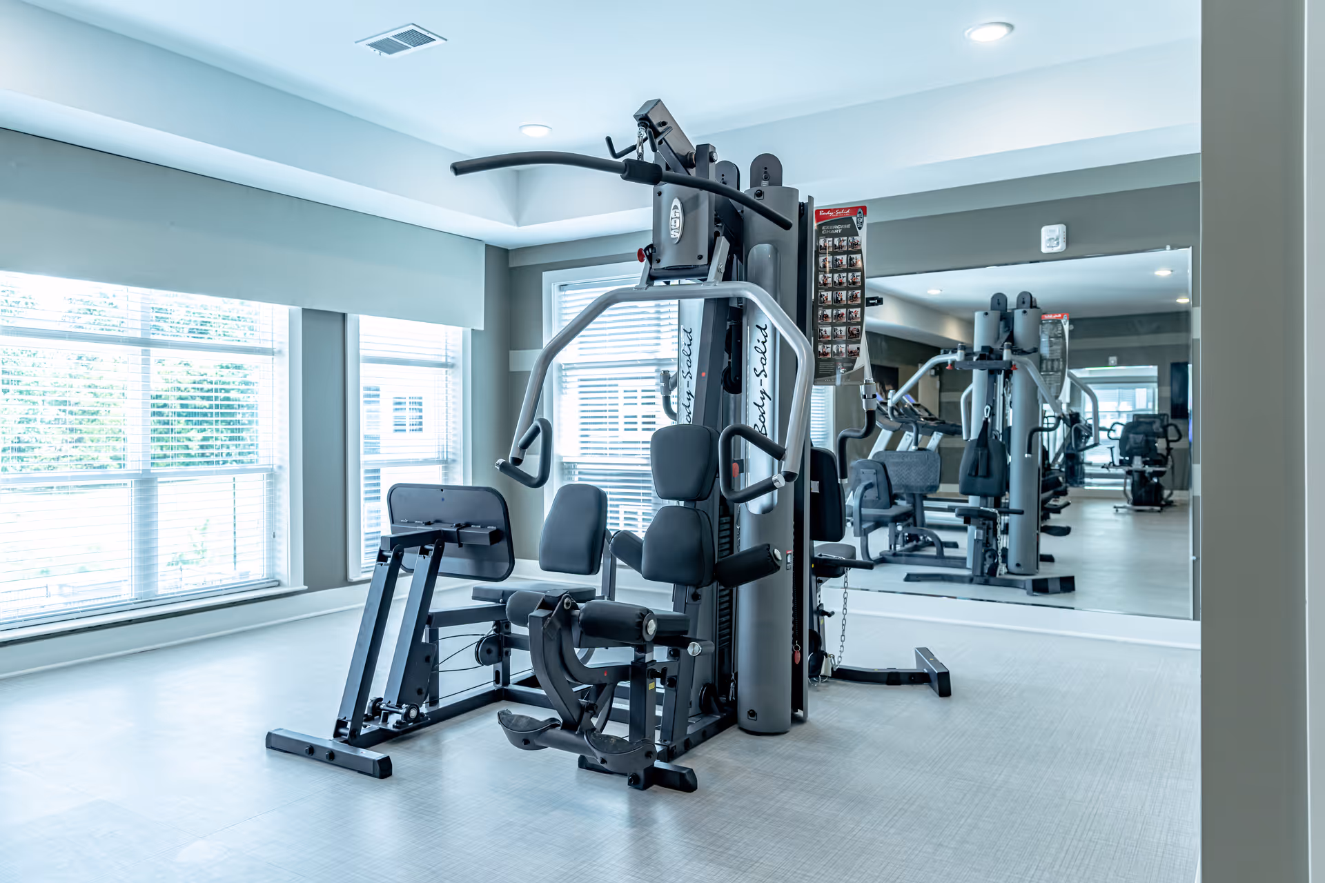 A bright fitness room with large windows allowing natural light to fill the space. The room features a multi-station gym machine with various exercise options, positioned in front of a large wall mirror reflecting the equipment and windows.