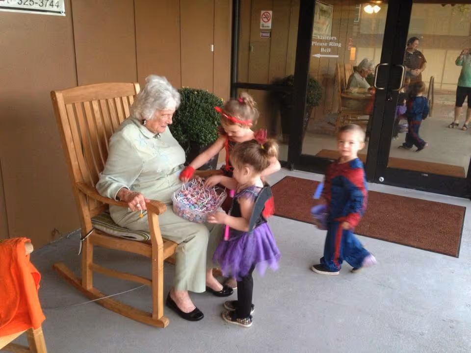 An elderly woman sitting in a wooden rocking chair outside a building, holding a bowl of candy while three young children in Halloween costumes gather around her. The children are dressed as superheroes and a fairy. The entrance door to the building is visible in the background with reflections of other people.