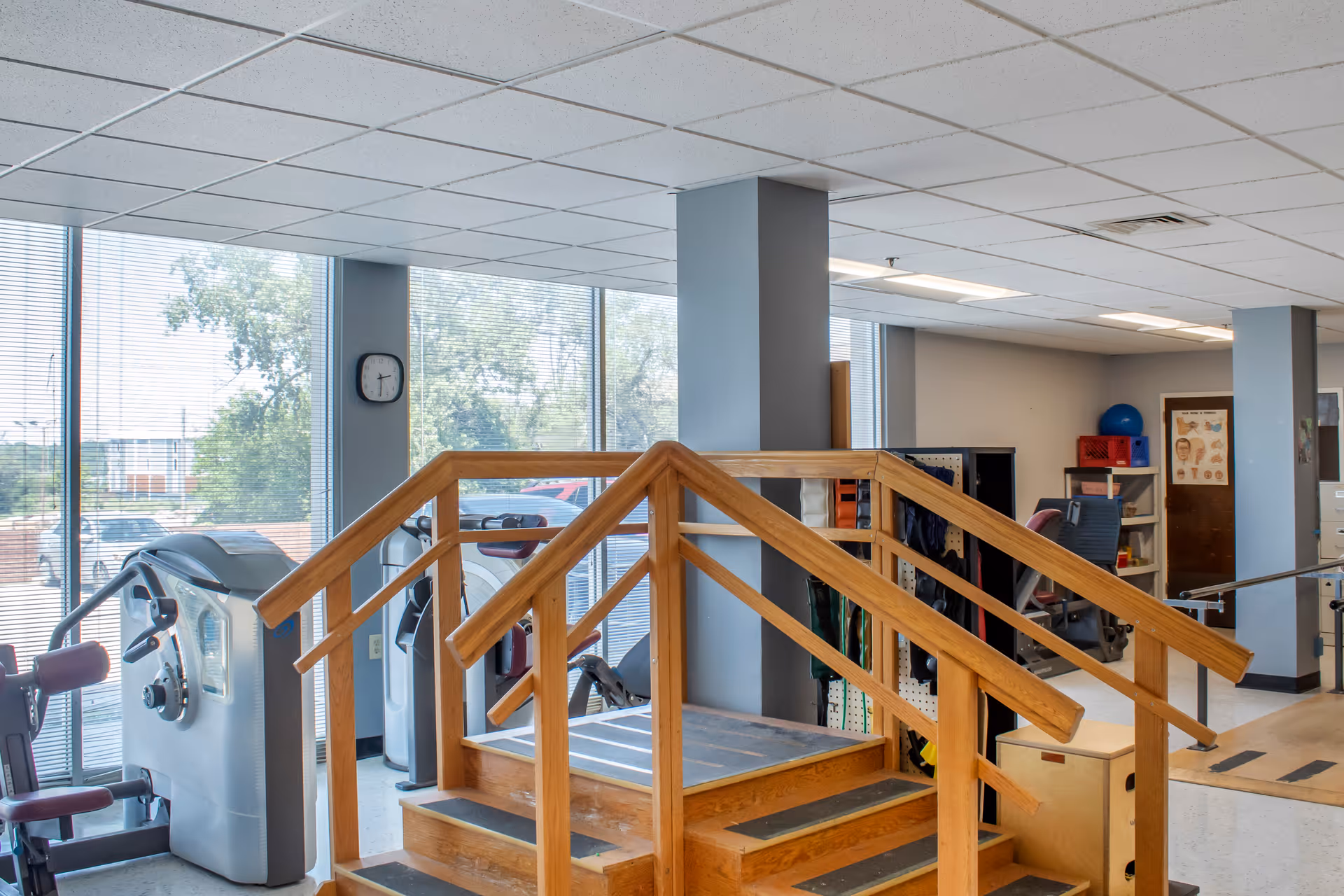 Interior view of a rehabilitation center room with exercise equipment, a wooden staircase with handrails for physical therapy, large windows with blinds, and various therapy tools and furniture in the background.
