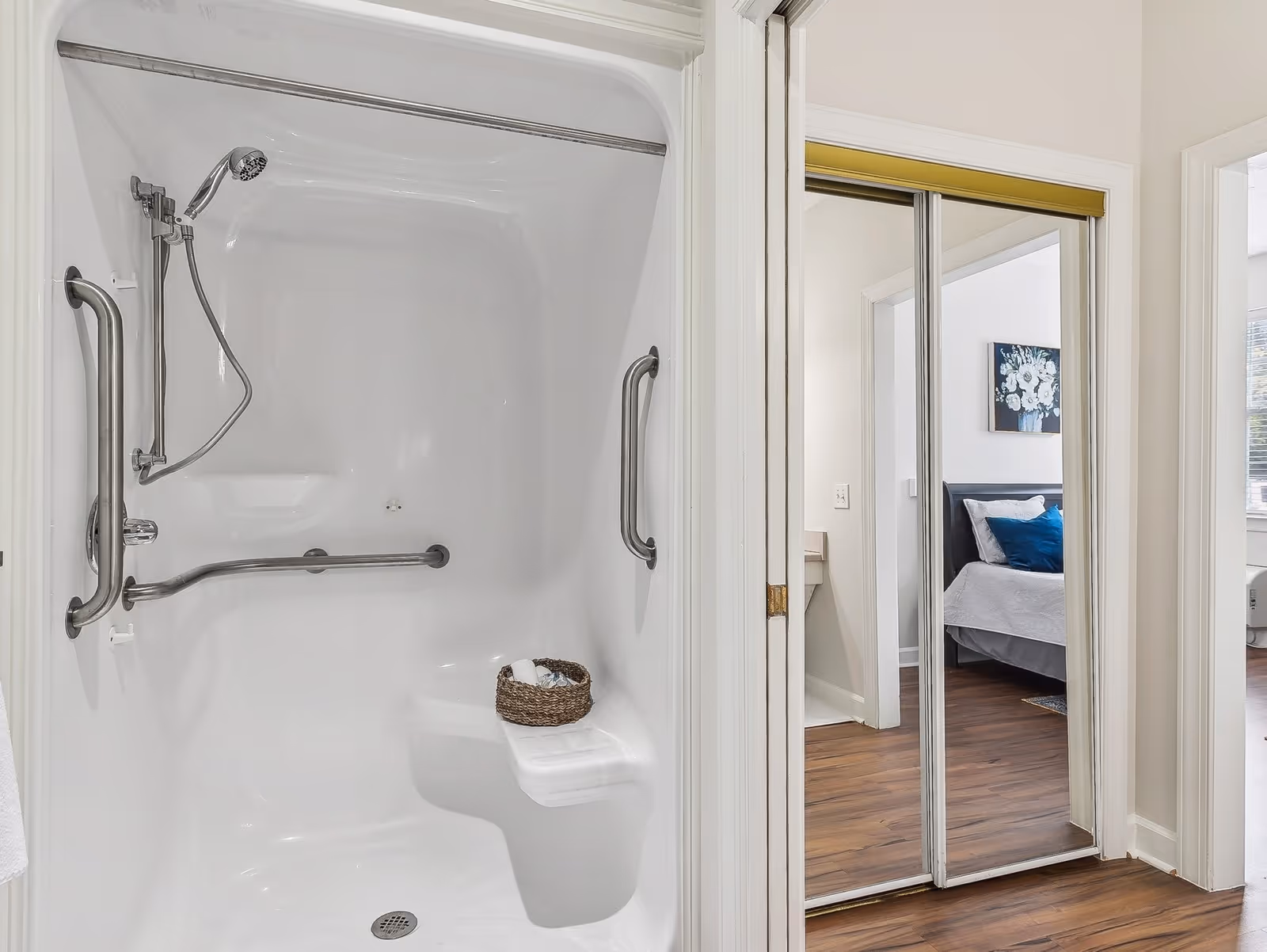 A white walk-in shower with built-in seat and multiple stainless steel grab bars. A handheld showerhead is mounted on the left wall. A small basket with toiletries sits on the seat. To the right, there is a doorway with mirrored sliding closet doors reflecting a bedroom with a bed, pillows, and a floral painting on the wall.
