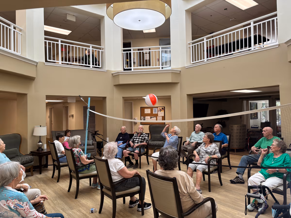 A group of elderly people seated in a circle of chairs playing a seated volleyball game with a beach ball indoors in a spacious room with high ceilings and a second-floor balcony.