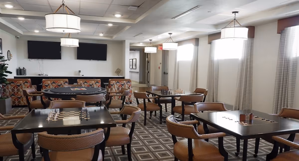 A well-lit game room with multiple tables and chairs arranged for playing board games such as chess and checkers. The room features patterned carpet, large windows with curtains, and modern hanging light fixtures. Two flat-screen TVs are mounted on the far wall above a seating area with colorful cushioned chairs.