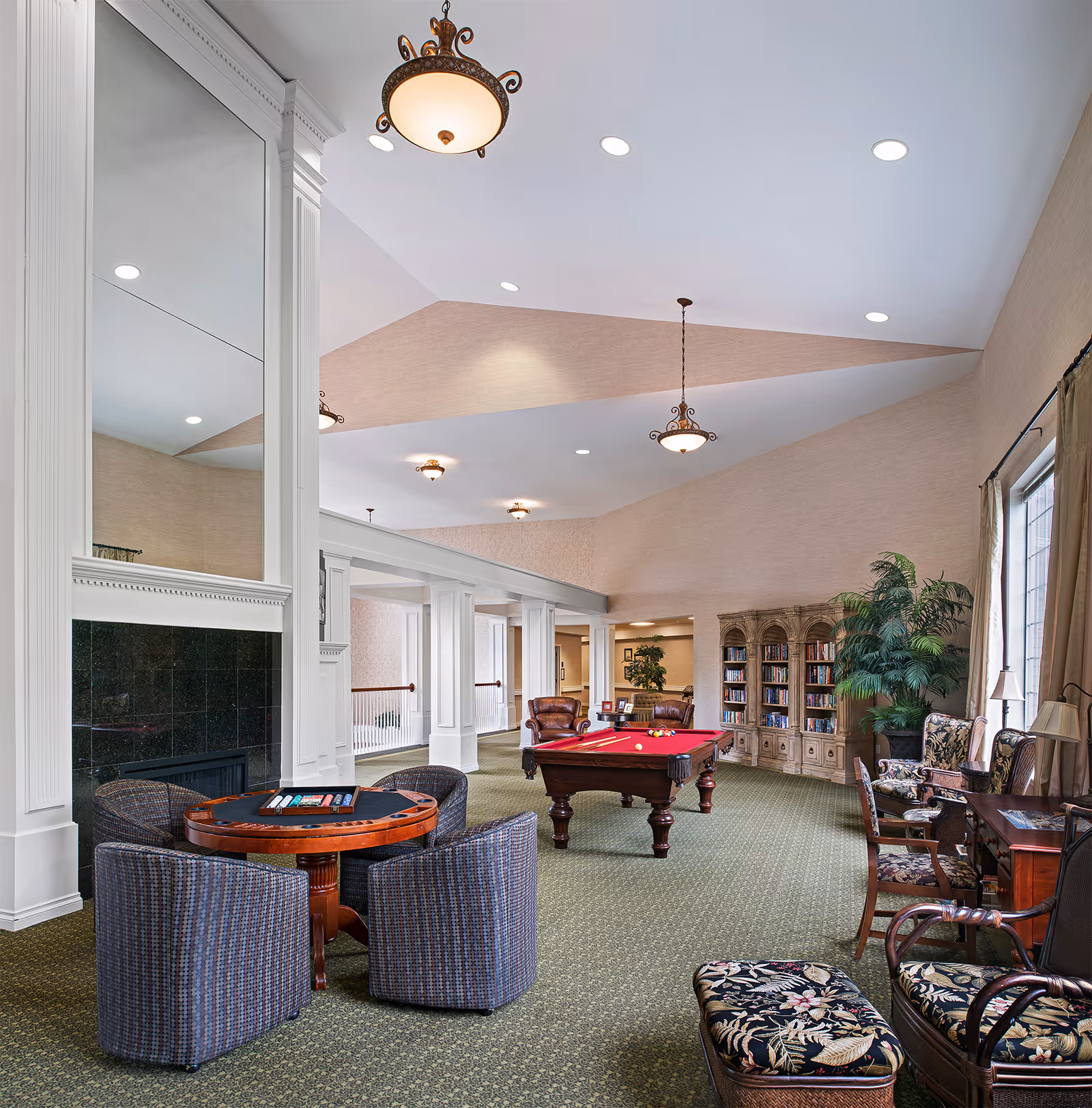 A spacious and well-lit common area in a senior living facility featuring a pool table with a red felt top, a round card table with four upholstered chairs, a large fireplace with a tall mirror above it, bookshelves filled with books, several armchairs with floral upholstery, and large windows with curtains allowing natural light to enter.