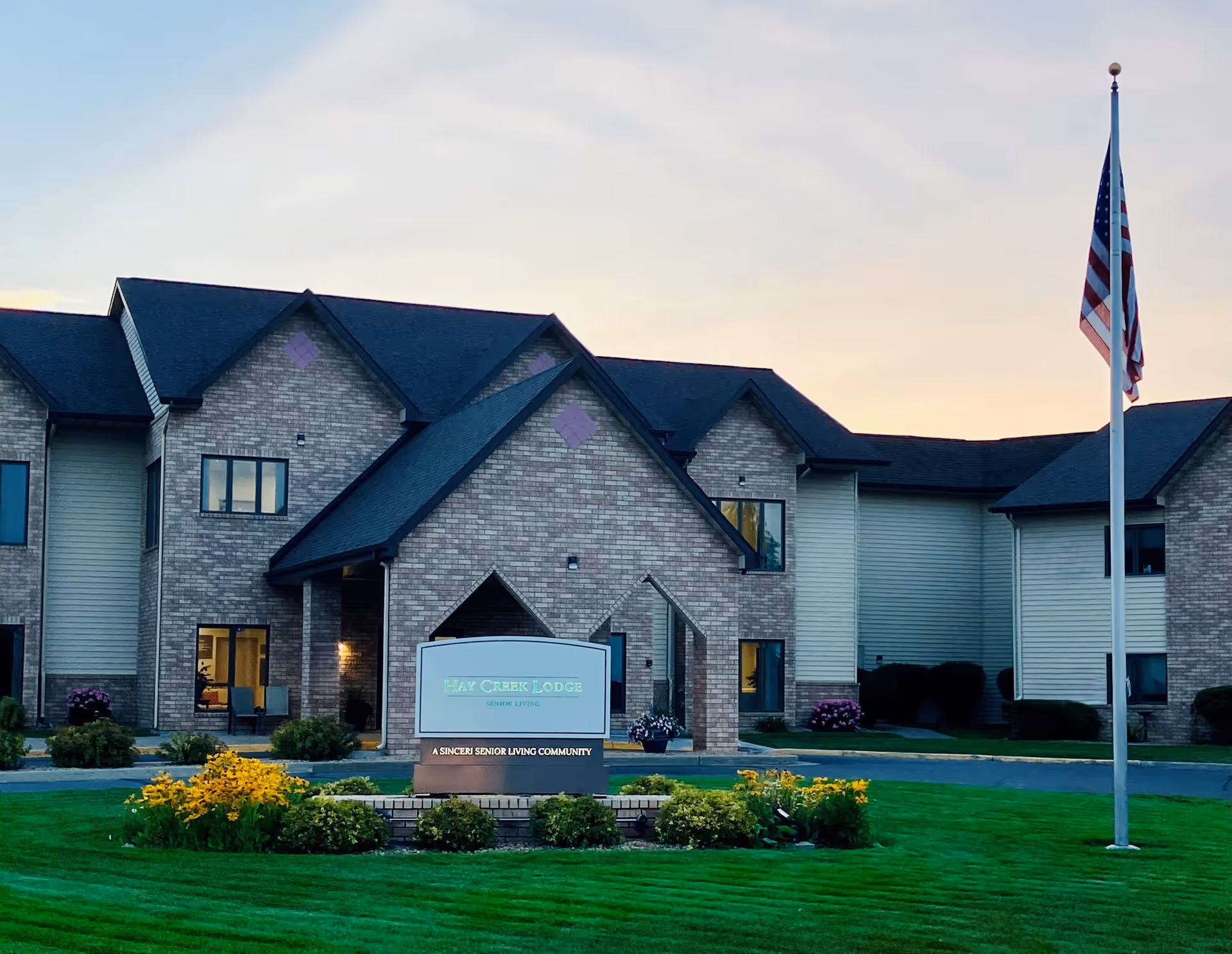 Exterior view of Hay Creek Lodge Senior Living building during sunset with a well-maintained lawn, flower beds, and an American flag on a flagpole in front.