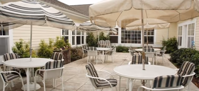 Outdoor patio area with multiple round white tables and striped cushioned chairs under large beige and gray umbrellas, surrounded by beige building walls and greenery.