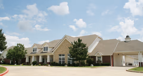 Exterior view of a single-story senior living facility building with a combination of brick and stone facade, multiple windows, a covered entrance, and a clear blue sky with some clouds above.