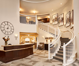 Spacious and elegant interior lobby area with a curved staircase featuring white railings and dark carpeted steps. The walls are decorated with framed artwork, and there is a large clock on the left wall above a dark wood reception desk with a flower arrangement. The area is well-lit with ceiling lights and wall sconces, and a seating area with a chair and lamps is visible near the staircase.