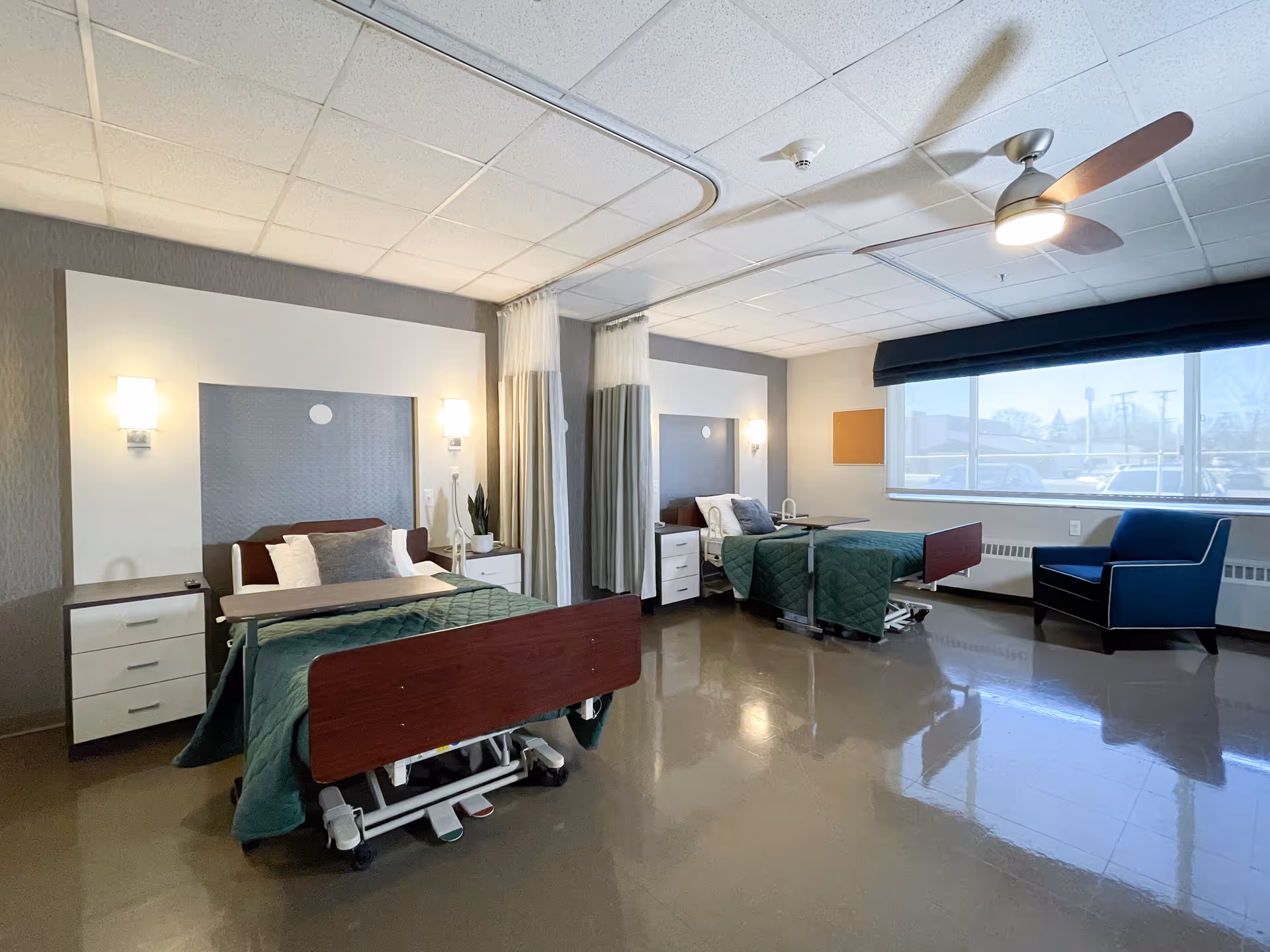 Spacious shared patient room with two adjustable beds, nightstands, seating area and a large window.