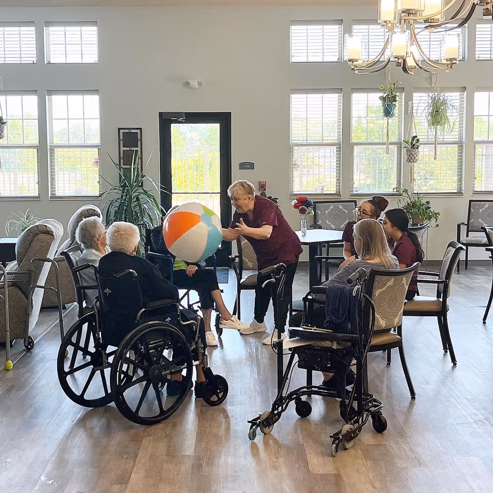 A group of elderly individuals and caregivers sitting in a bright, spacious room with large windows. Some elderly people are in wheelchairs, and a caregiver is playfully interacting with a large colorful beach ball. The room has wooden flooring, chairs, tables, and hanging plants near the windows.