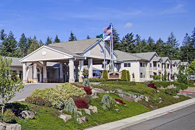 Exterior view of Harbor Place at Cottesmore, a senior living facility with a covered entrance, landscaped garden with shrubs and rocks, and two flagpoles displaying the American flag and another flag, surrounded by trees under a clear blue sky.