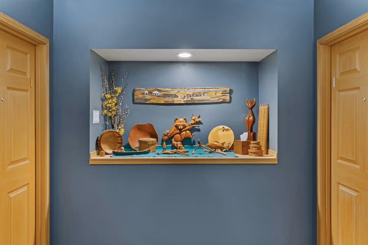 A recessed wall niche painted blue, displaying various wooden decorative items including a raccoon holding a branch, a cat figurine, plates, and other small wooden crafts, illuminated by a ceiling light. The niche is flanked by two wooden doors on either side.