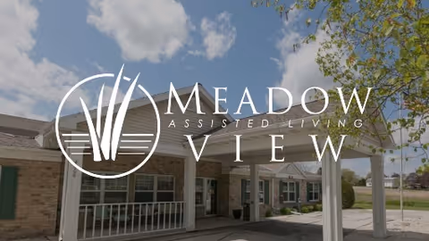 Exterior view of Meadow View Assisted Living facility showing a brick building with a covered entrance, white pillars, and a clear sky with some clouds.