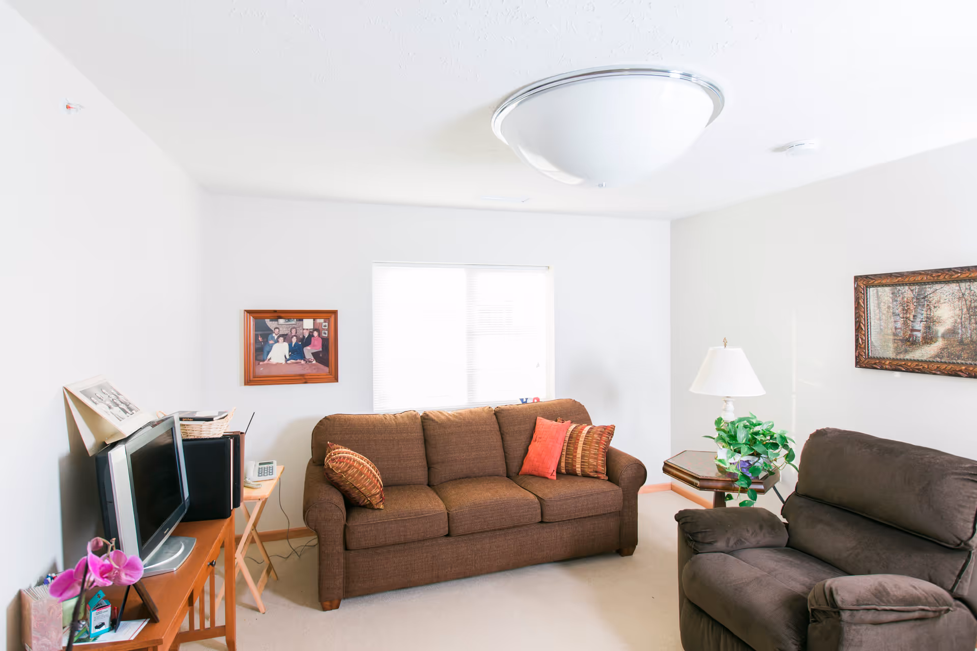 A cozy living room with a brown three-seater sofa adorned with three decorative pillows, a dark brown recliner chair, a wooden side table with a white lamp and a green potted plant, a wooden TV stand with a flat-screen TV and some framed photos, and two framed pictures hanging on the white walls.