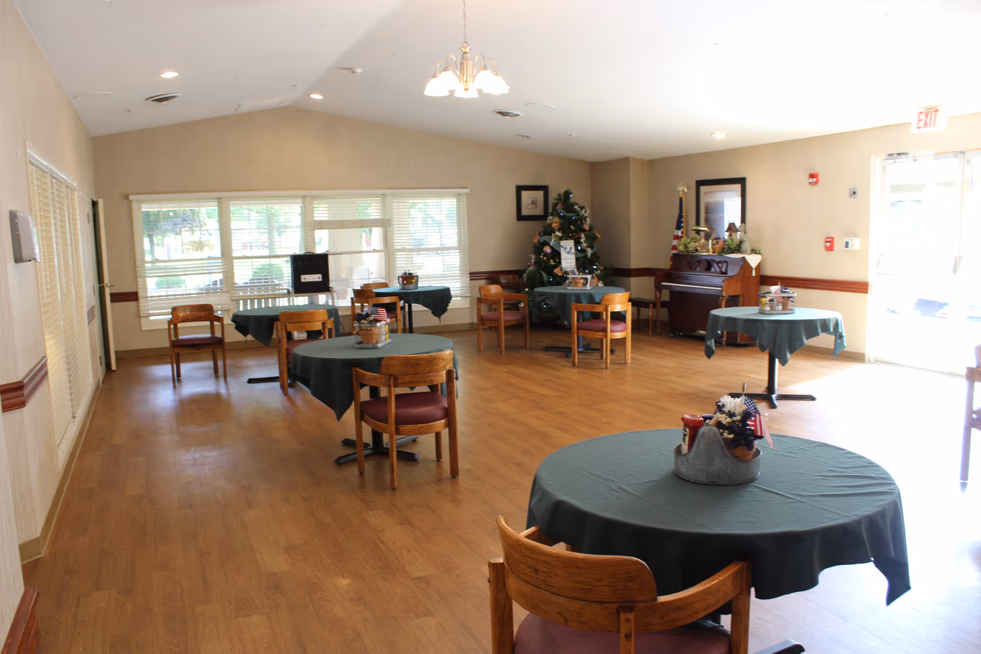 A bright dining room with wooden floors and several round tables covered with dark green tablecloths. Each table has a small centerpiece with flowers and decorative items. Wooden chairs with cushioned seats surround the tables. In the background, there is a decorated Christmas tree, a piano, and a large window letting in natural light. The room has beige walls and a ceiling light fixture.